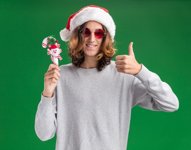 Heureux jeune homme portant chapeau de Père Noël et lunettes rouges tenant la canne de Noël regardant la caméra en souriant montrant les pouces vers le haut debout sur fond vert