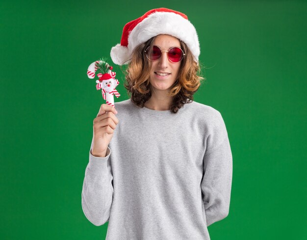Heureux jeune homme portant chapeau de Père Noël et lunettes rouges tenant la canne à sucre de Noël regardant la caméra avec le sourire sur le visage debout sur fond vert