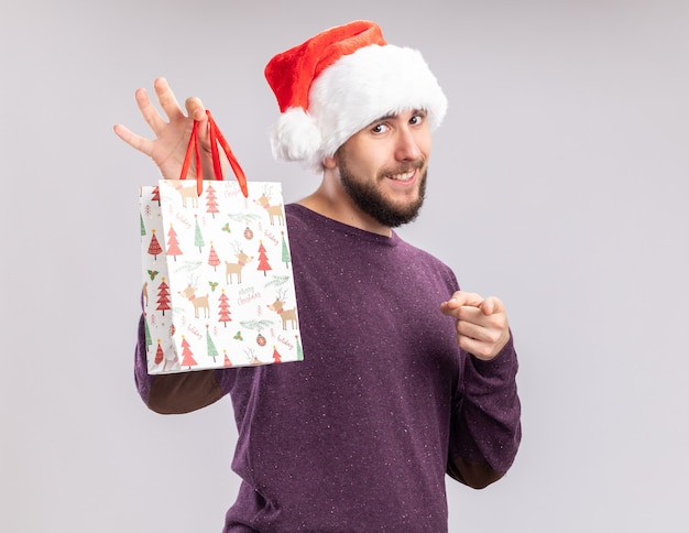 Heureux jeune homme en pull violet et bonnet de noel montrant un sac en papier cadeau pointant avec l'index à la caméra souriant joyeusement debout sur fond blanc