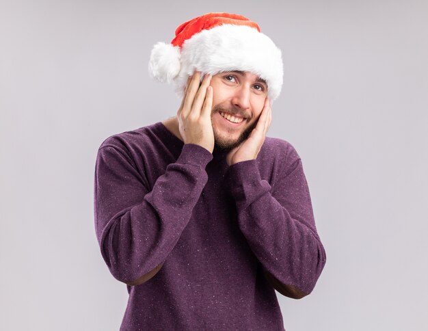 Heureux jeune homme en pull violet et bonnet de Noel regardant la caméra en souriant joyeusement avec les mains sur ses joues debout sur fond blanc