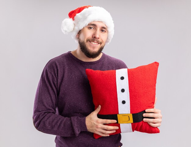 Heureux jeune homme en pull violet et bonnet de Noel tenant un oreiller drôle regardant la caméra avec le sourire sur le visage debout sur fond blanc