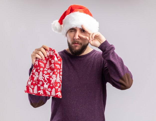 Heureux jeune homme en pull violet et bonnet de Noel tenant un sac rouge avec des cadeaux montrant v-signe sur son œil debout sur fond blanc