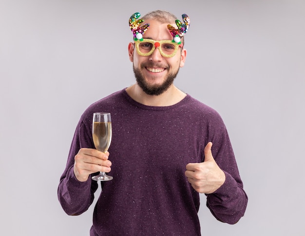 Heureux jeune homme en pull violet et lunettes drôles tenant un verre de champagne regardant la caméra avec le sourire sur le visage ferrant les pouces vers le haut debout sur fond blanc