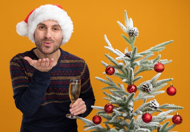 Heureux jeune homme de race blanche portant un chapeau de Noël debout près de sapin de Noël décoré tenant un verre de champagne regardant la caméra envoi baiser coup isolé sur fond orange