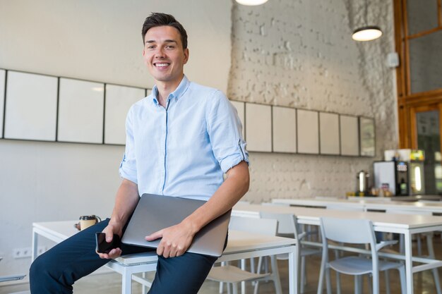 Heureux jeune homme souriant attrayant assis dans un bureau ouvert de co-working, tenant un ordinateur portable,
