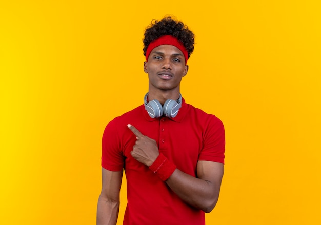 Heureux jeune homme sportif afro-américain portant bandeau et bracelet avec des points d'écouteurs sur le côté isolé sur fond jaune avec copie espace