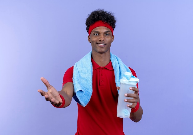 Heureux jeune homme sportif afro-américain portant un bandeau et un bracelet tenant une bouteille d'eau tendant la main à la caméra avec une serviette sur l'épaule