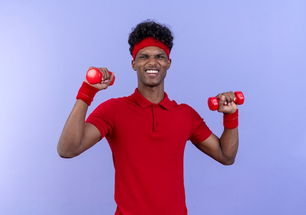 Heureux jeune homme sportif afro-américain portant bandeau et bracelet tenant haltère isolé sur mur bleu