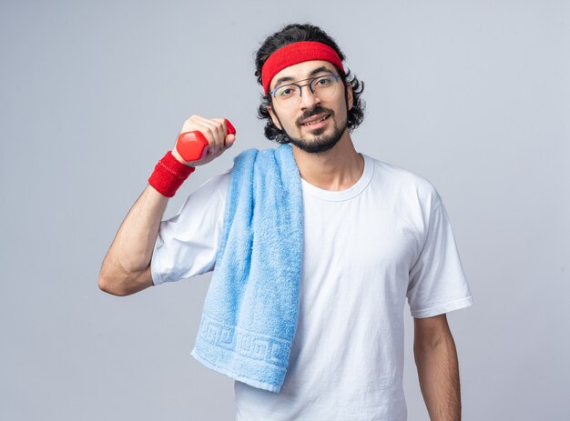 Heureux jeune homme sportif portant un bandeau avec un bracelet et une serviette sur l'épaule faisant de l'exercice avec un haltère