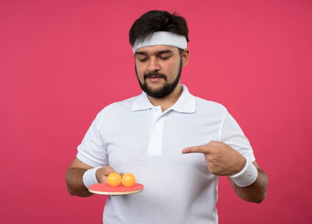 Heureux jeune homme sportif portant un bandeau et un bracelet tenant et des points à une raquette de ping-pong avec des balles