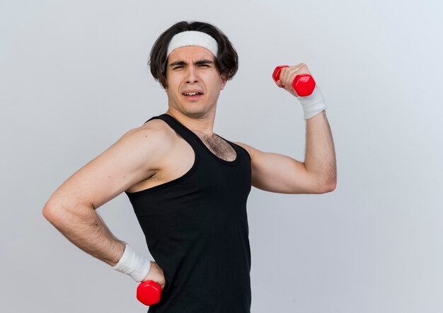 Heureux jeune homme sportif portant des vêtements de sport et un bandeau travaillant avec des haltères souriant posant à l'avant debout sur un mur blanc