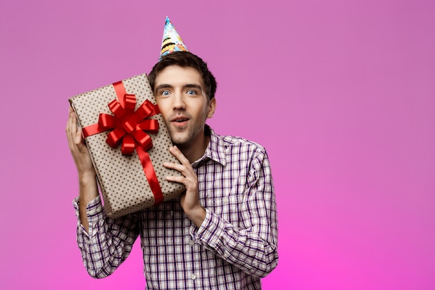 Heureux jeune homme tenant un cadeau d'anniversaire dans une boîte sur le mur violet.