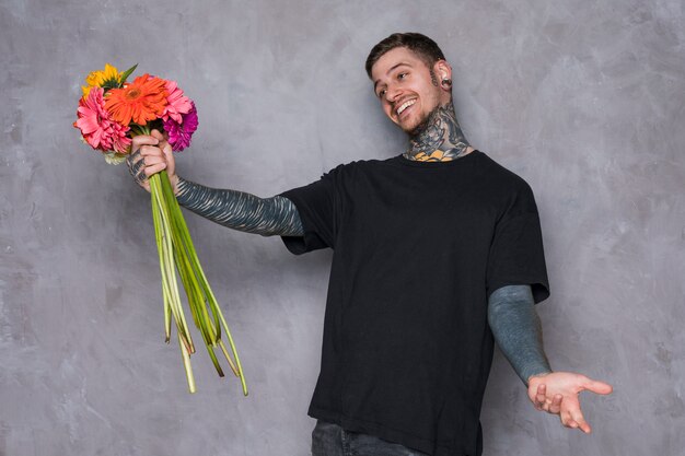 Heureux jeune homme tenant des fleurs de gerbera en main haussant les épaules contre le mur gris