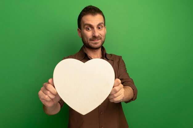 Heureux jeune homme tenant et regardant en forme de coeur isolé sur mur vert