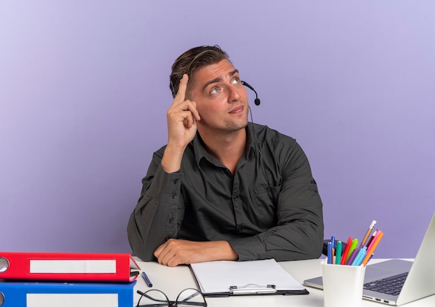 Heureux jeune homme de travailleur de bureau blonde au casque est assis au bureau avec des outils de bureau à l'aide de points d'ordinateur portable et lève les yeux