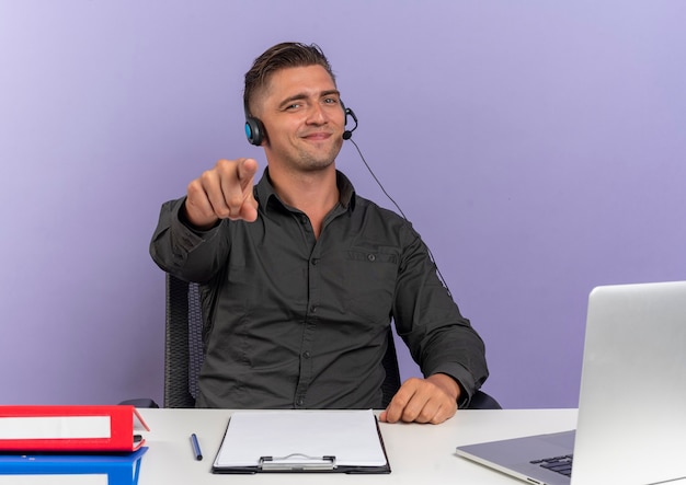 Heureux jeune homme travailleur de bureau blonde sur le casque est assis au bureau avec des outils de bureau à l'aide de points d'ordinateur portable à la caméra isolée sur fond violet avec espace de copie