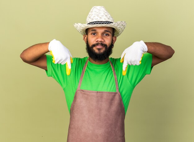 Heureux jeune jardinier afro-américain portant un chapeau de jardinage avec des gants pointe vers le bas