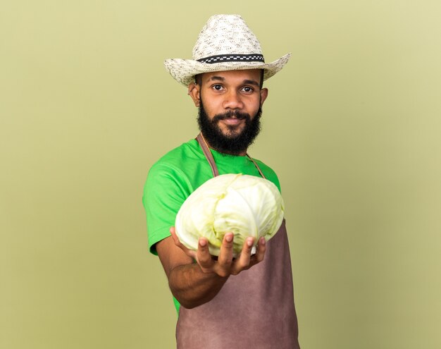 Heureux jeune jardinier afro-américain portant un chapeau de jardinage tenant du chou à la caméra