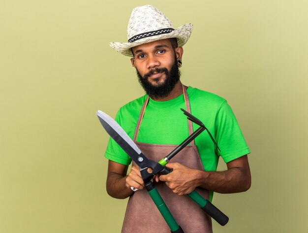 Heureux jeune jardinier afro-américain portant un chapeau de jardinage tenant et traversant des tondeuses avec un râteau à houe isolé sur un mur vert olive