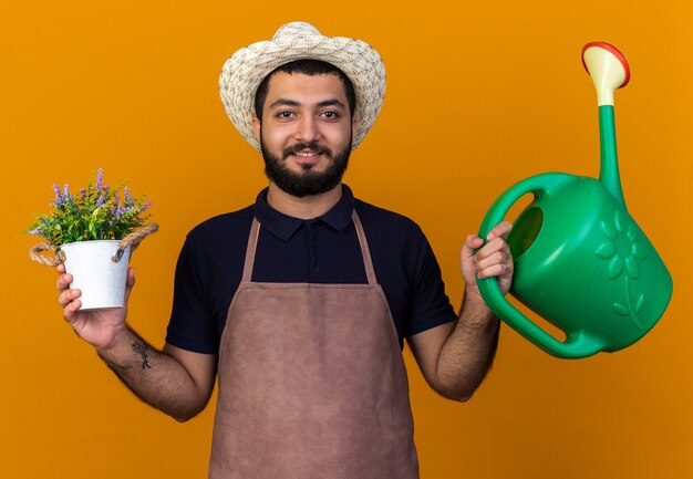 Heureux jeune jardinier caucasien portant un chapeau de jardinage tenant un arrosoir et un pot de fleurs isolé sur un mur orange avec espace pour copie