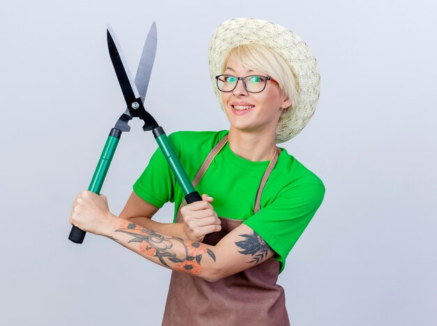 Heureux jeune jardinier femme aux cheveux courts en tablier et chapeau montrant des coupe-haies souriant gaiement