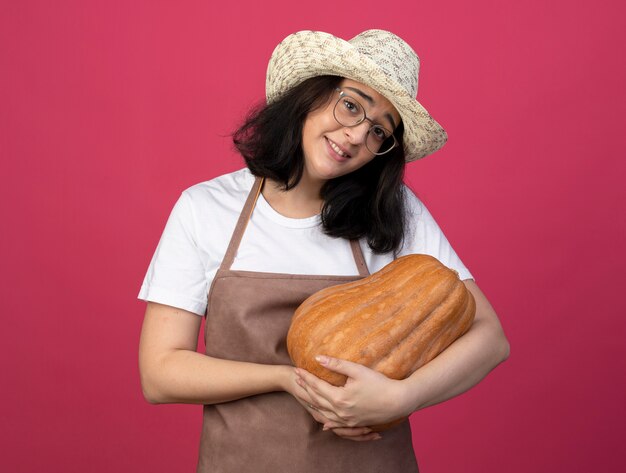 Heureux jeune jardinier femme brune à lunettes optiques et en uniforme portant chapeau de jardinage détient citrouille isolé sur mur rose avec espace copie