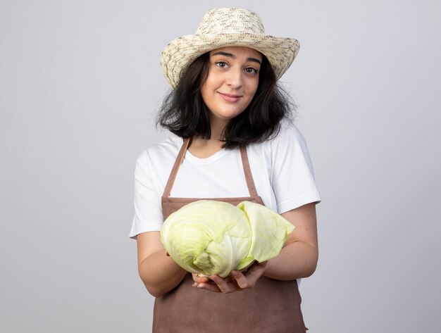 Heureux jeune jardinier femme brune en uniforme portant chapeau de jardinage détient chou isolé sur mur blanc