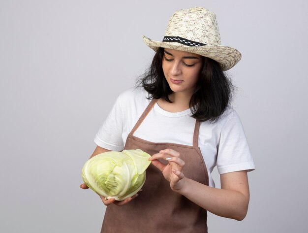 Heureux jeune jardinier femme brune en uniforme portant chapeau de jardinage tient et regarde le chou isolé sur un mur blanc avec copie espace