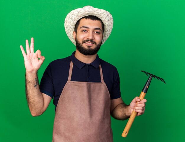 Heureux jeune jardinier mâle caucasien portant chapeau de jardinage tenant râteau et gesticulant signe ok isolé sur mur vert avec espace copie