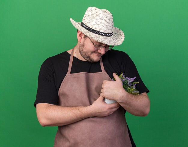 Heureux jeune jardinier mâle portant chapeau de jardinage attrapé et regardant la fleur en pot de fleurs isolé sur mur vert