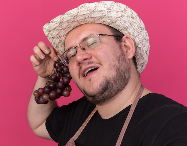 Heureux jeune jardinier mâle portant chapeau de jardinage tenant des raisins et avant isolé sur mur rose