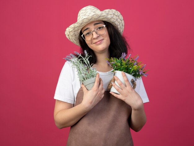 Heureux jeune jardinière brune à lunettes optiques et en uniforme portant chapeau de jardinage détient des pots de fleurs isolés sur un mur rose avec copie espace