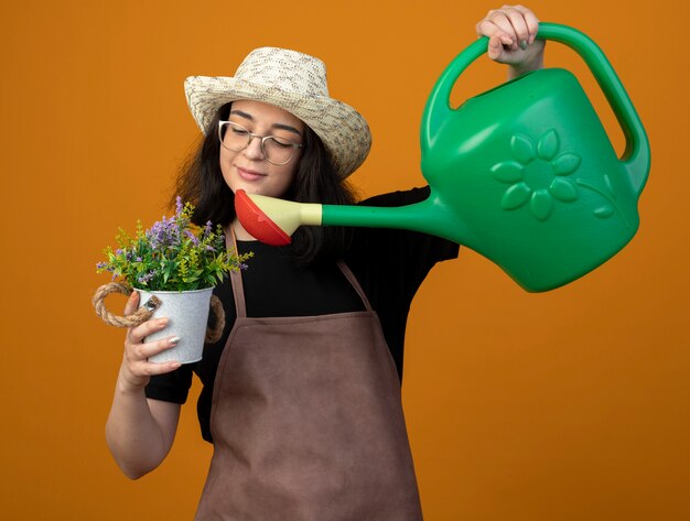 Heureux jeune jardinière brune à lunettes optiques et en uniforme portant un chapeau de jardinage fait semblant d'arroser les fleurs en pot de fleurs avec arrosoir isolé sur mur orange avec espace de copie