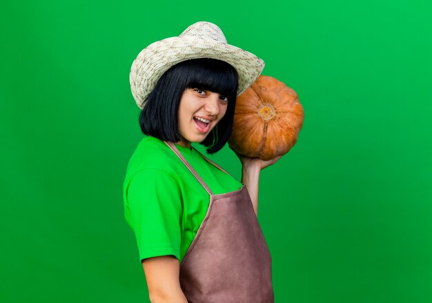 Heureux jeune jardinière en uniforme portant un chapeau de jardinage détient citrouille