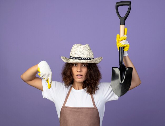 Heureux jeune jardinière en uniforme portant chapeau de jardinage et points de gants avec pelle à bas