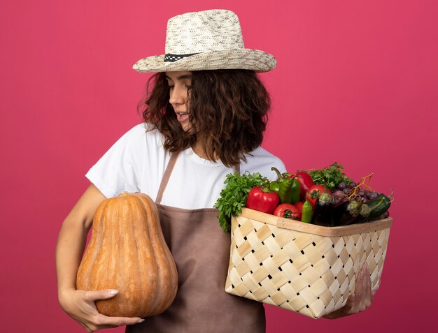 Photo gratuite heureux jeune jardinière en uniforme portant chapeau de jardinage tenant un panier de légumes et regardant la citrouille dans sa main isolé sur rose