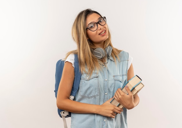 Heureux jeune jolie étudiante portant des lunettes et sac à dos avec des écouteurs sur le cou tenant des livres isolés sur un mur blanc