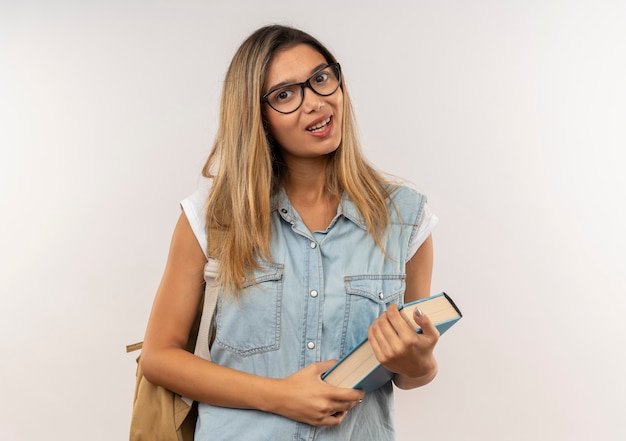 Heureux jeune jolie étudiante portant des lunettes et sac à dos tenant livre isolé sur mur blanc