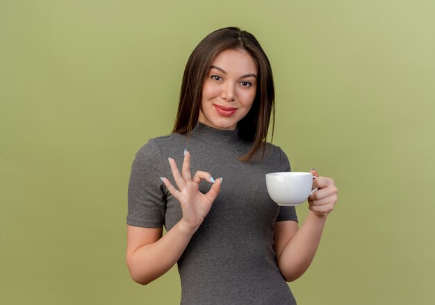 Heureux jeune jolie femme tenant la tasse et faisant signe ok isolé sur fond vert olive avec espace copie