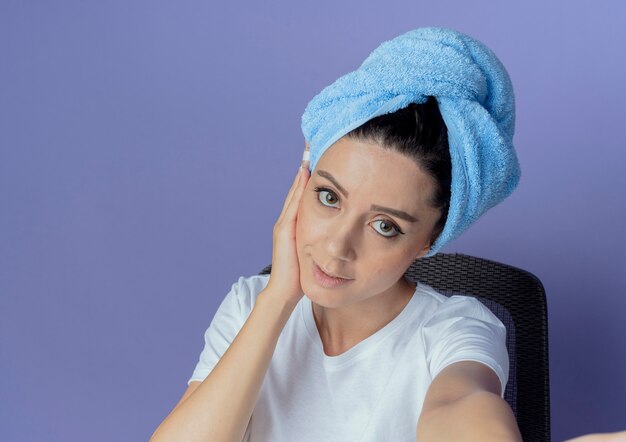 Heureux jeune jolie fille assise à la table de maquillage avec des outils de maquillage et avec une serviette de bain sur la tête mettant la main sur le visage isolé sur fond violet avec espace copie