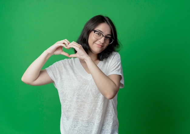 Heureux jeune jolie fille caucasienne portant des lunettes faisant signe de coeur isolé sur fond vert avec espace copie