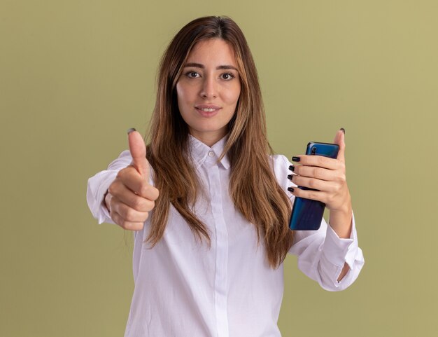 Heureux jeune jolie fille caucasienne pouces vers le haut et tient le téléphone isolé sur un mur vert olive avec espace de copie