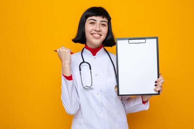 Heureux jeune jolie fille caucasienne en uniforme de médecin avec stéthoscope tenant un stylo et un presse-papiers
