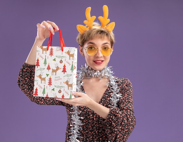 Heureux jeune jolie fille portant bandeau de bois de renne et guirlande de guirlandes autour du cou avec des lunettes tenant le sac-cadeau de Noël regardant la caméra isolée sur fond violet avec espace de copie