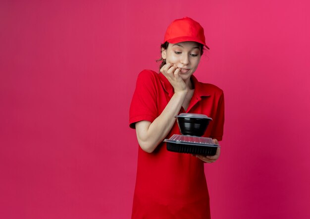 Heureux jeune jolie livreuse portant uniforme rouge et cap tenant et regardant les contenants de nourriture avec la main sur le menton isolé sur fond cramoisi avec espace de copie