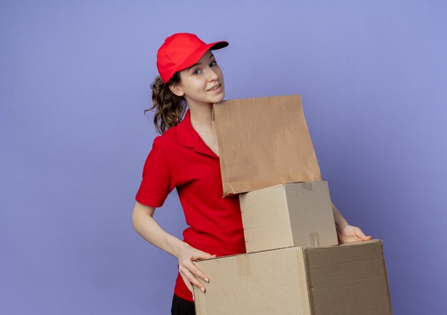 Heureux jeune jolie livreuse portant un uniforme rouge et une casquette tenant des boîtes en carton et un paquet de papier isolé sur fond violet avec espace de copie