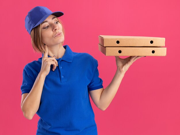Heureux jeune jolie livreuse en uniforme met le doigt sur le menton tenant et regardant des boîtes de pizza sur rose