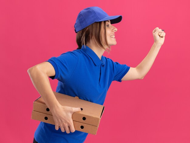 Heureux jeune jolie livreuse en uniforme se tient sur le côté tenant des boîtes de pizza faisant semblant de courir sur le rose