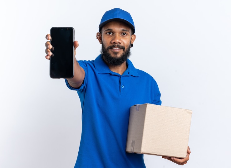 Photo gratuit heureux jeune livreur afro-américain tenant une boîte en carton et un téléphone isolé sur fond blanc avec espace pour copie