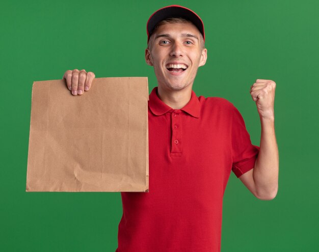 Heureux jeune livreur blond garde le poing et tient le paquet de papier